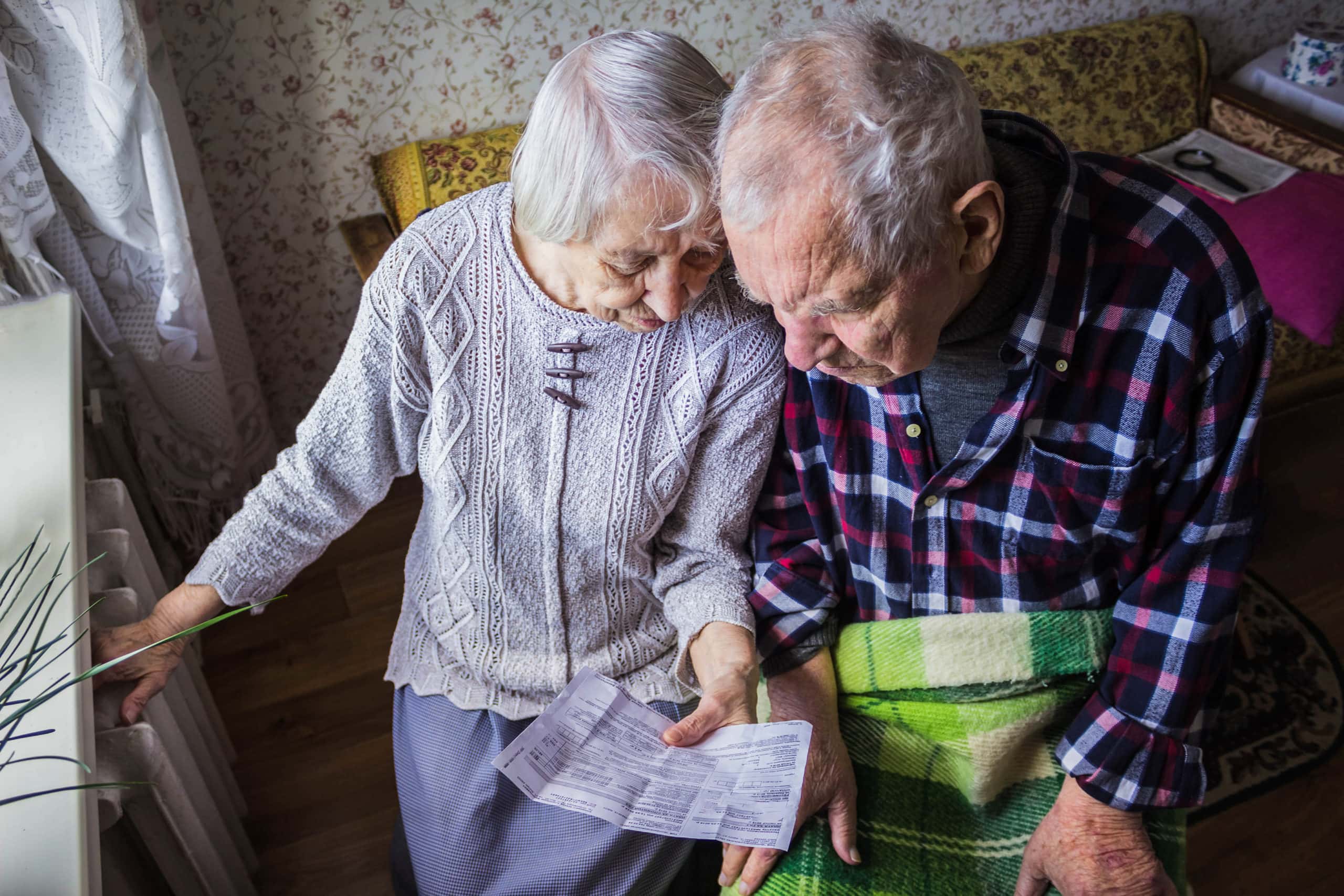 The senior woman and man holding gas bill in front of heating radiator. Payment for heating in winter.