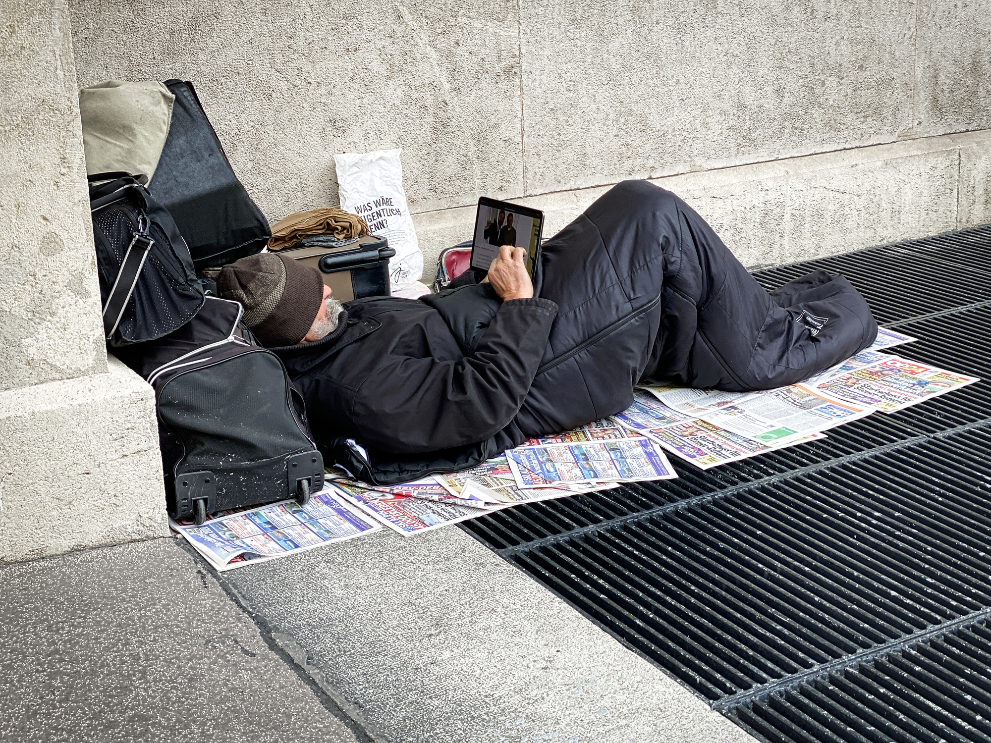 Vienna, Austria - January 30, 2020: Homeless man laying on warm ventilation grill and look internet on his tablet