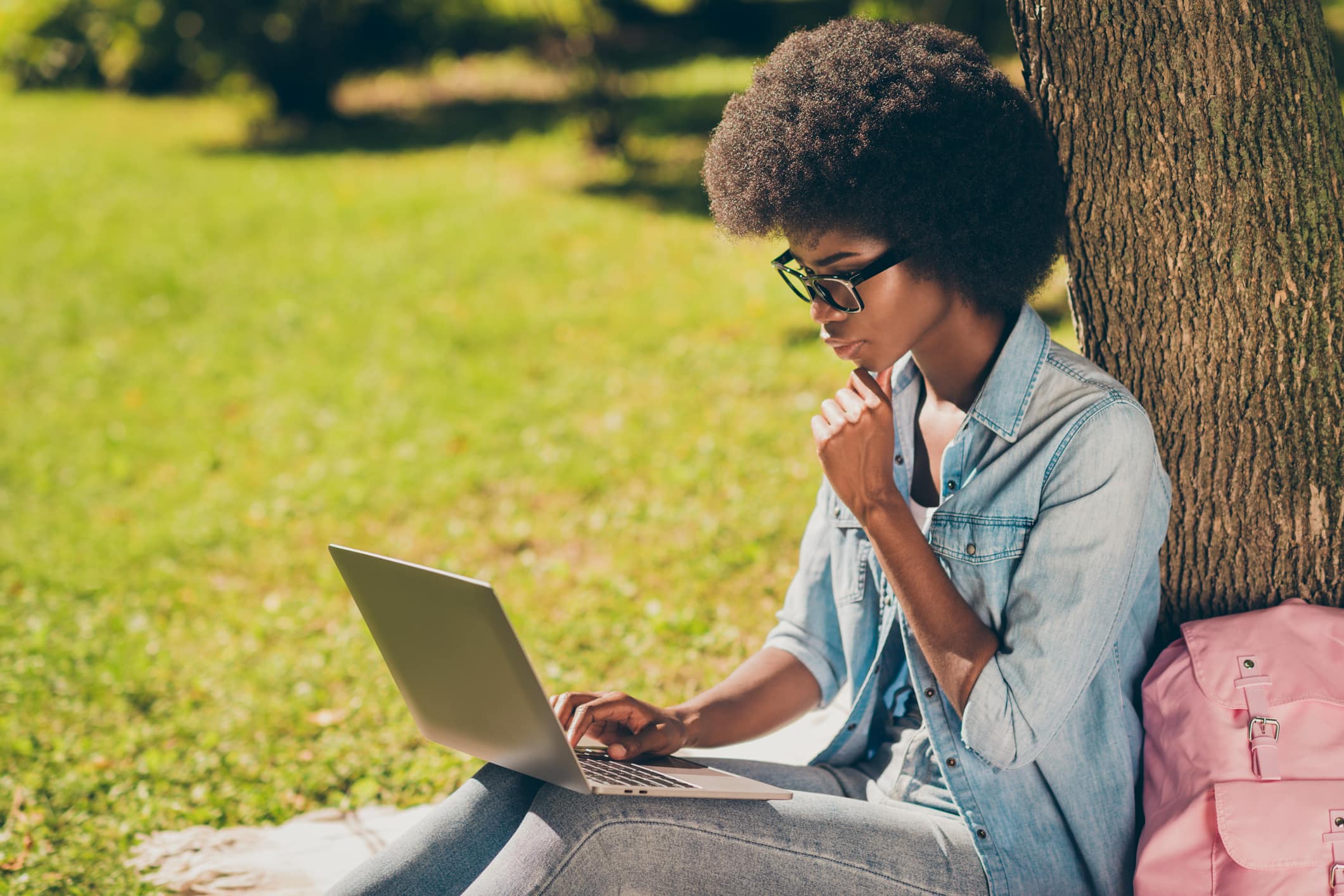 Side profile photo of black skinned serious girl using laptop touching chin learning outside the city park wearing eyewear.